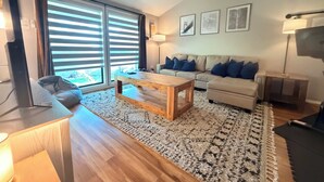 A modern living room with a wooden coffee table, a sofa with blue cushions, a large window with blinds, and a flat-screen TV. - Sleek Urban Retreat in Happy Valley (Happy Valley)