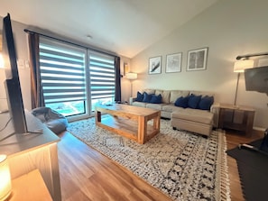A modern living room with a wooden coffee table, a sofa with blue cushions, a large window with blinds, and a flat-screen TV. - Sleek Urban Retreat in Happy Valley (Happy Valley)