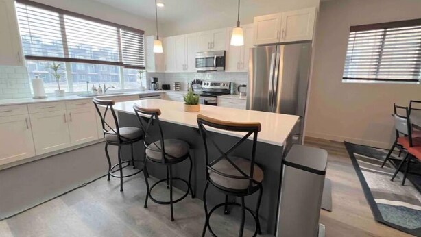 A modern kitchen with white cabinets, a central island with bar stools, stainless steel appliances, and large windows with blinds. - Modern Townhouse w/ All Kings and Private Rooftop (West Valley City)
