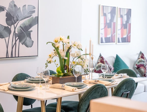 A dining area with a table set for four, featuring a centerpiece of white orchids, candles, and a wooden box. There are green upholstered chairs and a white sofa in the background.