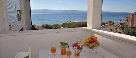 Una mesa en el balcón dispuesta con platos, cubiertos, vasos de jugo de naranja, un frutero y un florero con flores.