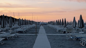 A beach with rows of sun loungers and umbrellas, a central walkway, and a sunset in the background.