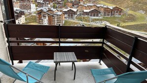 En balkong med en träbänk och två blå stolar, med utsikt över en stad med trähus och en fri vy över det omgivande landskapet.