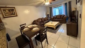 Una sala de estar con una mesa de comedor preparada para cuatro personas, un sofá, un televisor y un ventilador de techo.