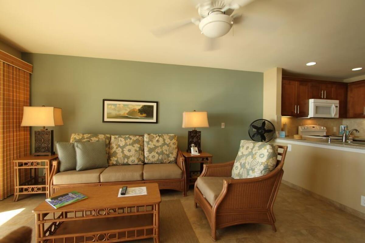 A living room with a sofa, two side tables with lamps, a wicker chair, a ceiling fan, and a kitchen area with a microwave.