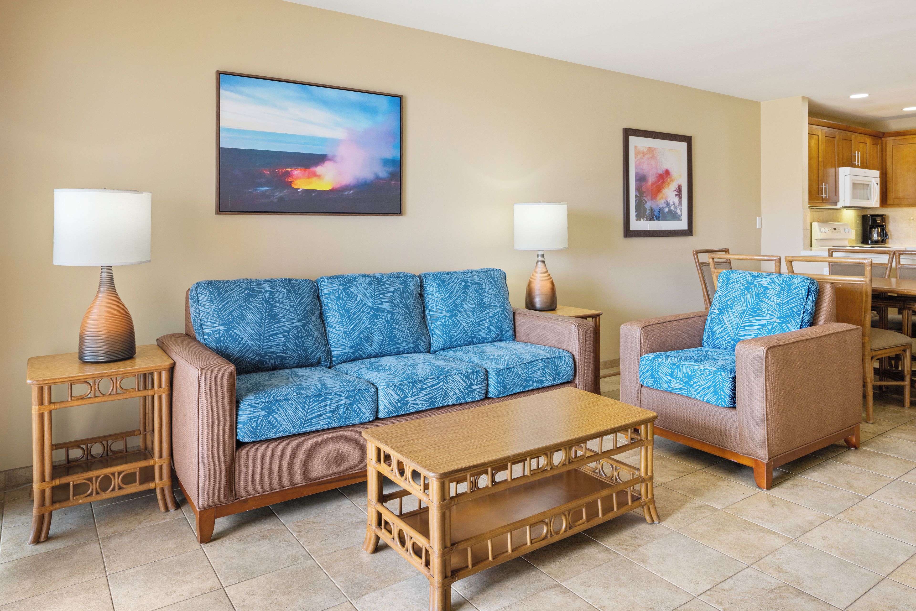 A living room with a sofa, armchairs, a coffee table, and lamps. There is a kitchen in the background.