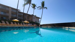 A clear blue pool with lounge chairs and umbrellas, surrounded by a building with balconies and palm trees. - Sea Village (Kailua-Kona)