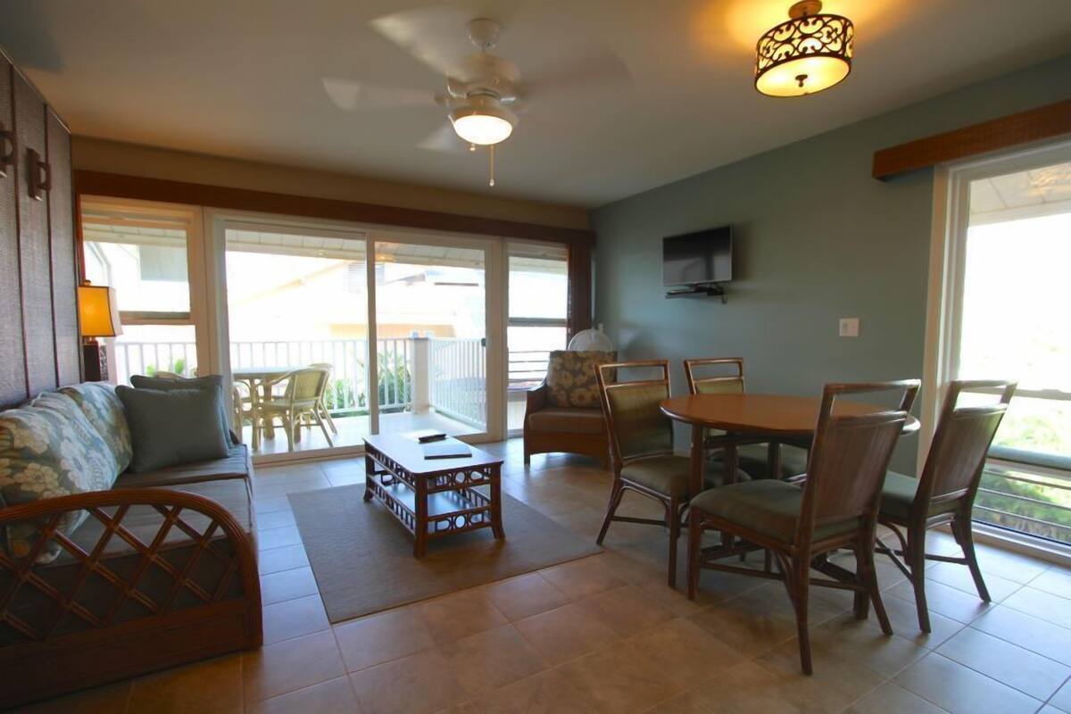 A spacious living area with a sofa, dining table, and chairs, a ceiling fan, and a TV mounted on the wall.