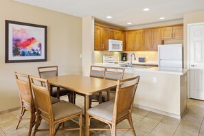 A modern kitchen with wooden cabinets, a white refrigerator, and a dining area with a table and chairs. - Sea Village (Kailua-Kona)