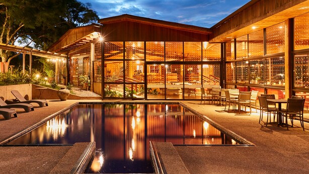 A pool area with a wooden pergola, outdoor seating, and a wine cellar visible through large windows.