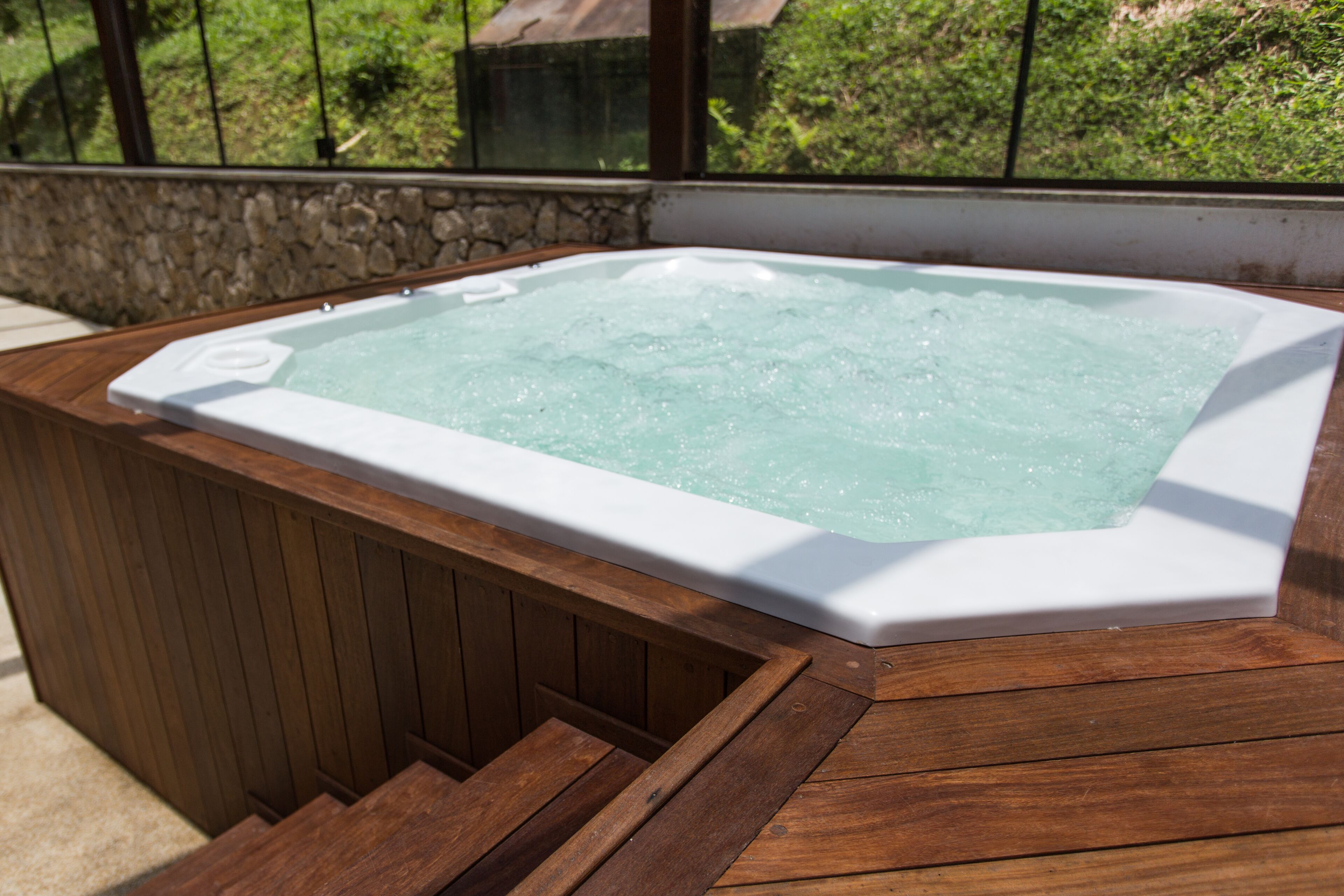A hot tub with a wooden deck and stone wall backdrop.