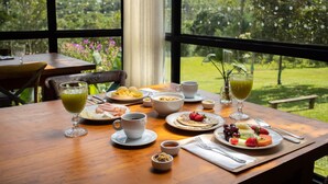 A breakfast table with various dishes, including pancakes, fruit, and a green drink, set against a backdrop of a lush garden and a large window.