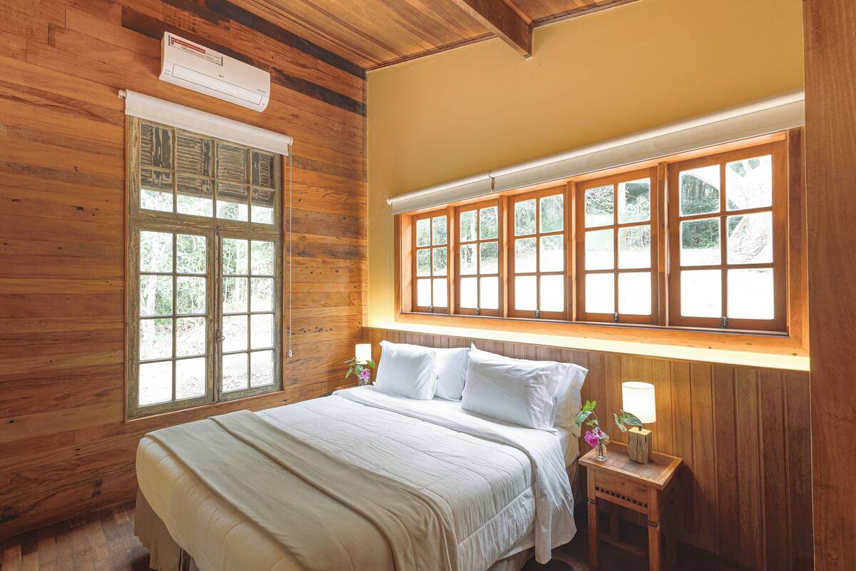 A wooden-paneled room with a large bed, a window with shutters, and a small bedside table with a lamp.