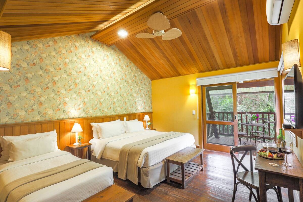 A room with two beds, wooden ceiling, floral wallpaper, and a balcony with outdoor furniture.