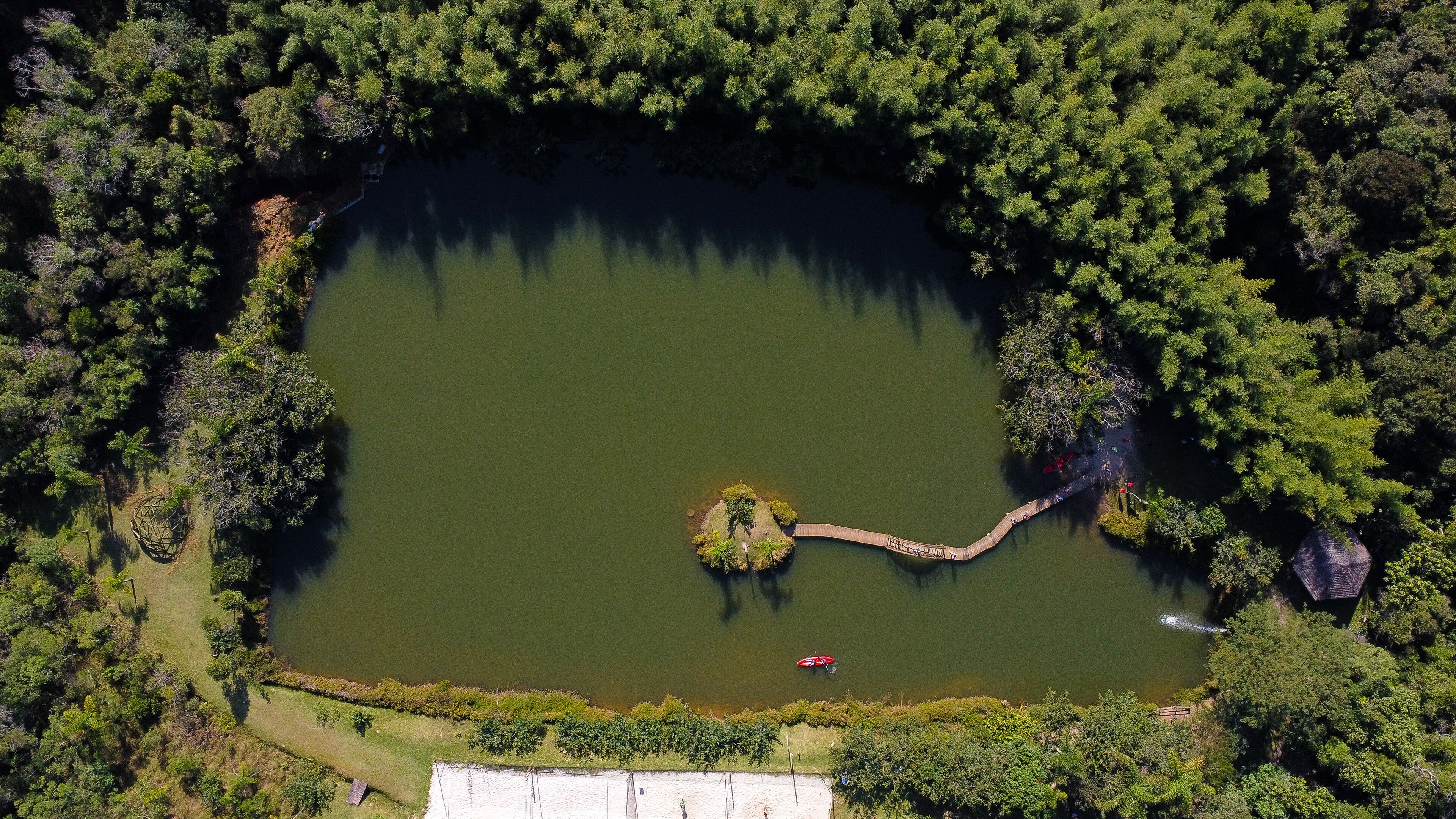 Vista aérea de um lago rodeado por floresta densa, com uma pequena ilha no centro ligada à margem por uma ponte estreita.