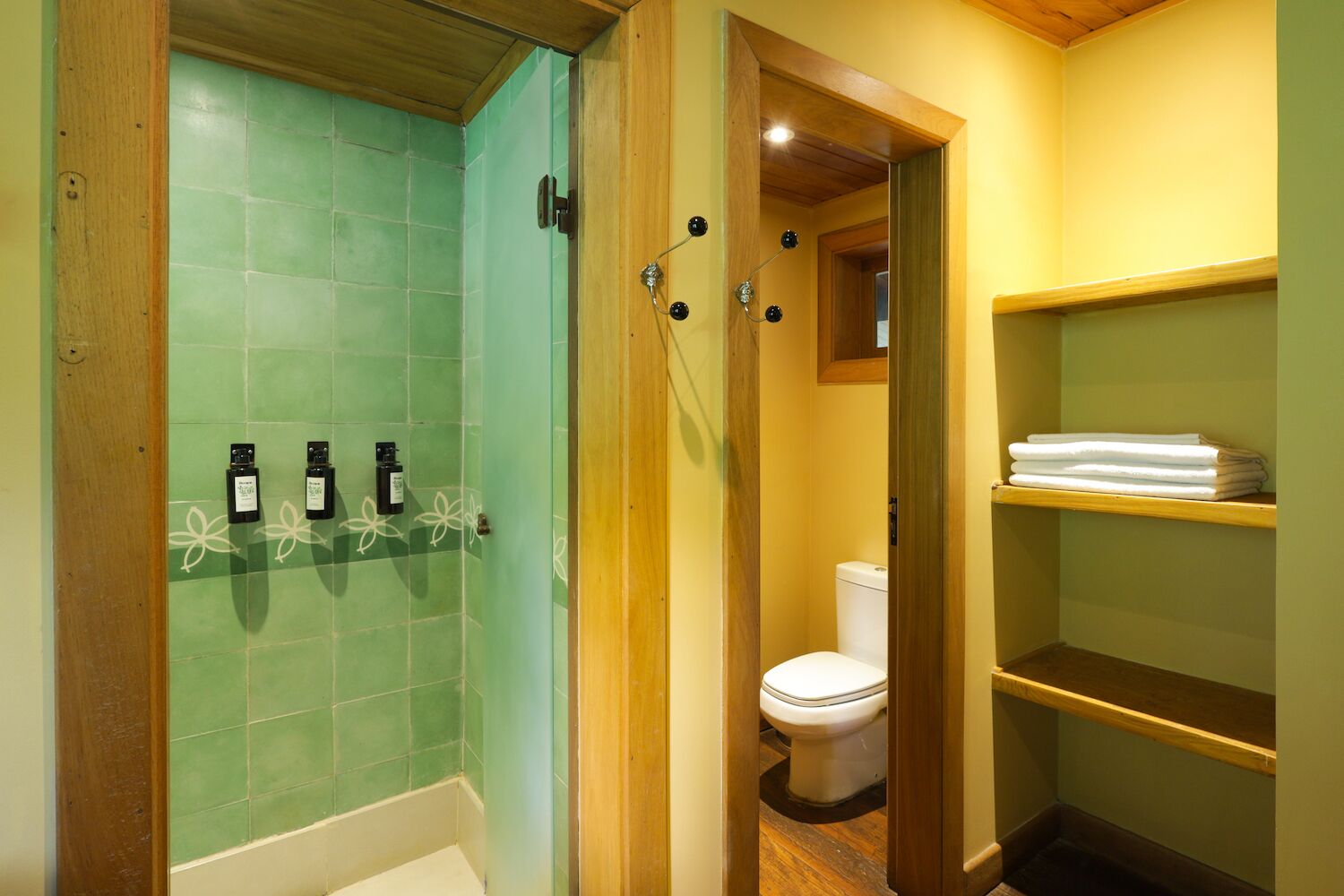 Un baño con ducha de azulejos verdes, un inodoro blanco y estantes de madera con toallas dobladas.
