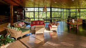 A spacious indoor area with wooden flooring and furniture, large windows offering a view of greenery outside.