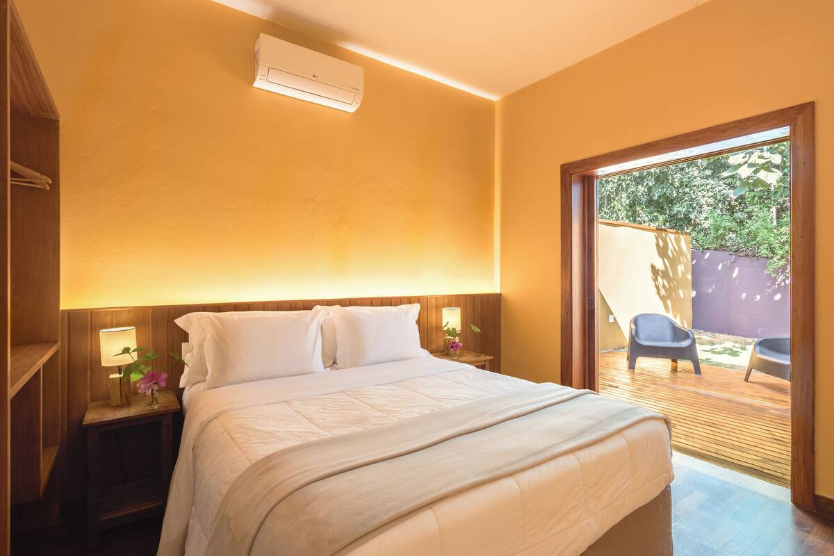 A bedroom with a bed, bedside tables, an air conditioning unit, and a view of an outdoor area with chairs.