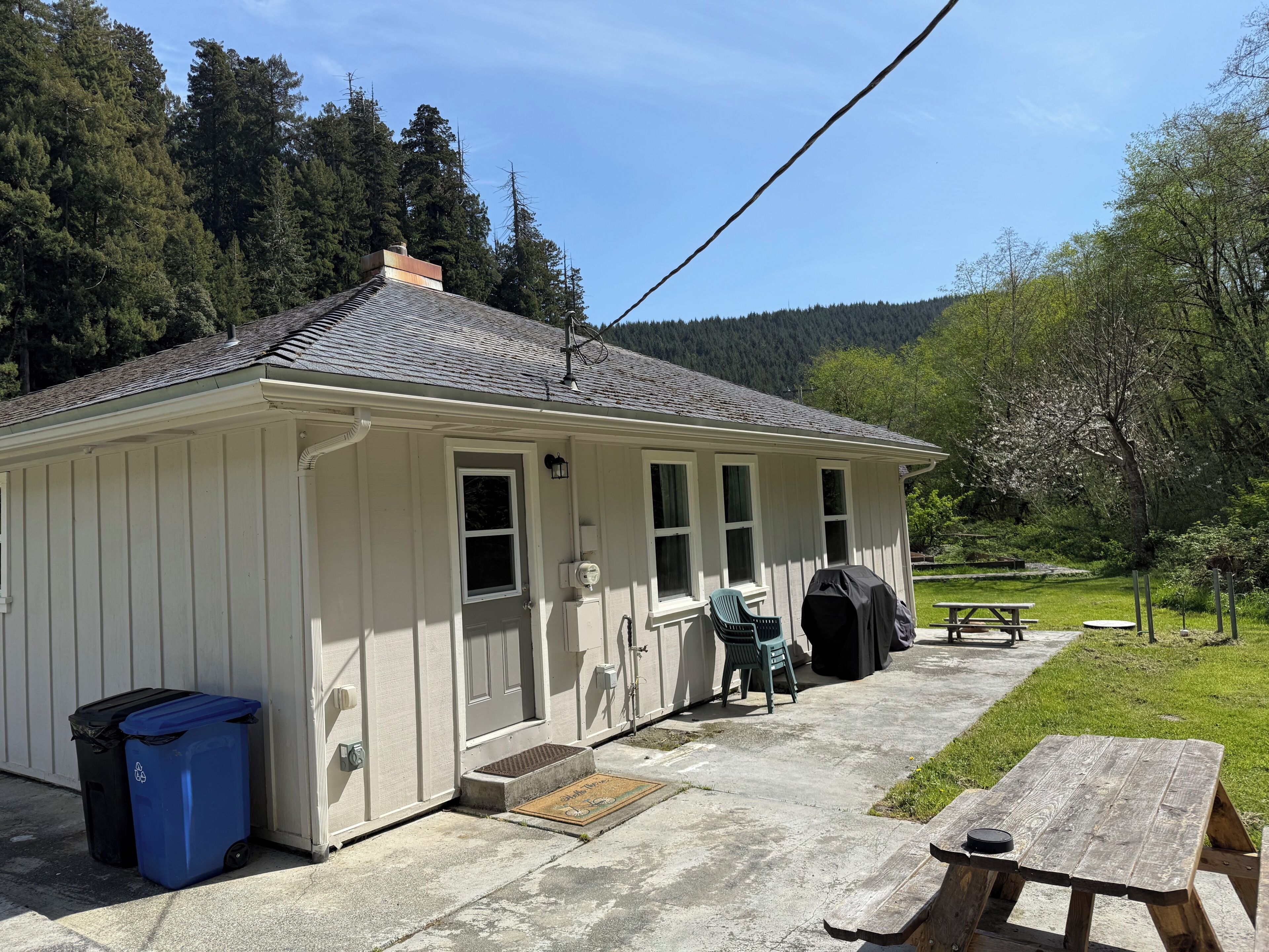 A small, single-story building with a porch, a barbecue grill, and a picnic table.