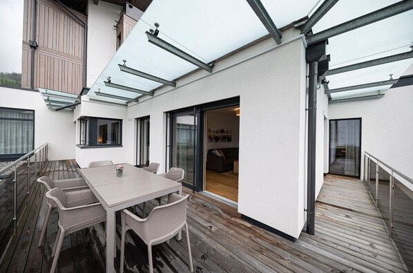 A modern outdoor patio with a wooden deck, a light-colored table with chairs, and a glass door leading to an interior space.
