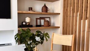 Una habitación con estantes de madera que exhiben varios objetos, una silla, una planta en maceta y una mesa redonda de madera.