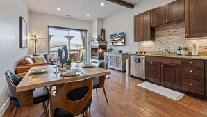 A modern kitchen with wooden cabinets, a dining area with a wooden table and chairs, and a large window offering a view of the city.