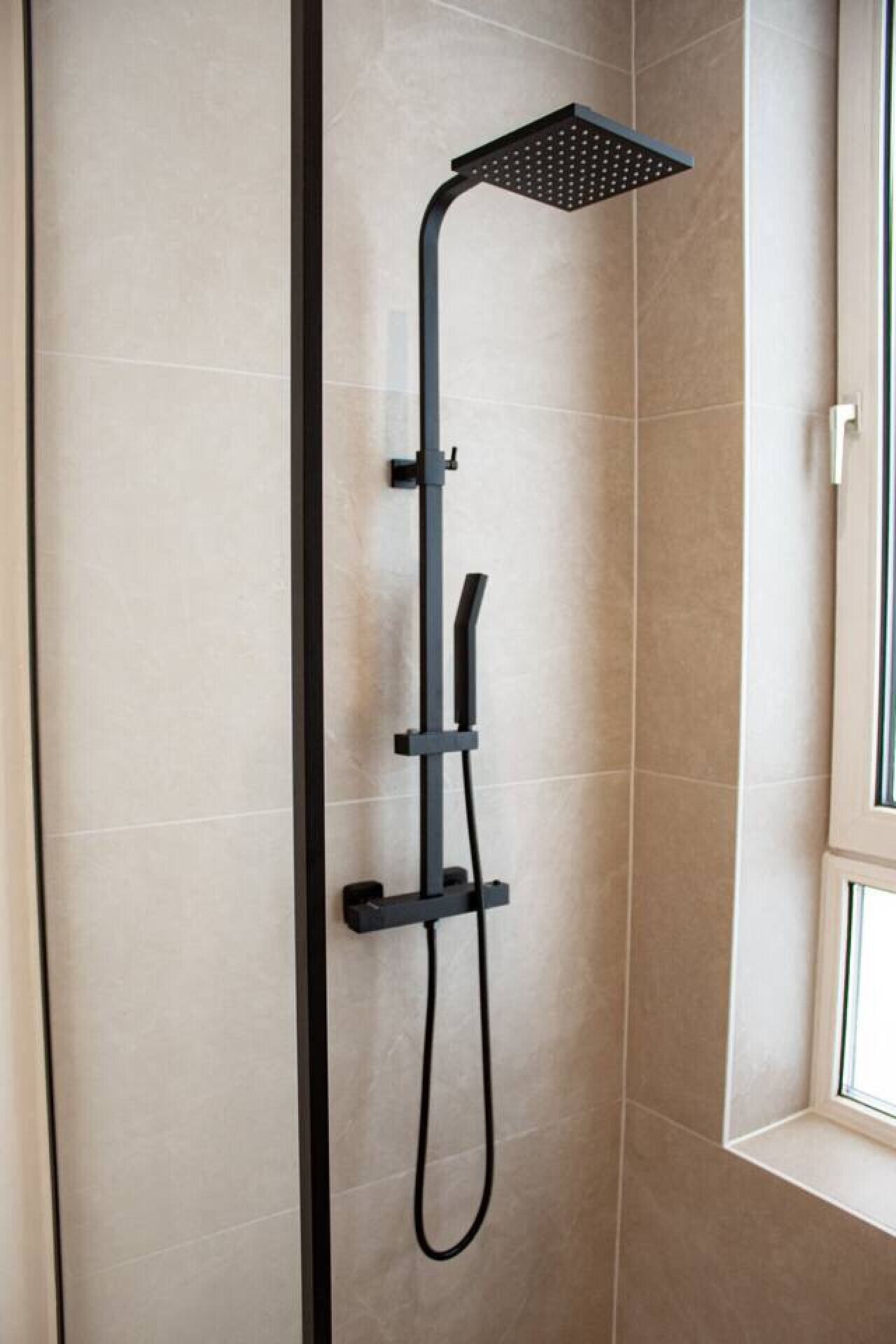 Une douche moderne dotée d’un pommeau de douche carré et d’un pommeau de douche à main, fixée sur un mur avec une fenêtre sur le côté droit.