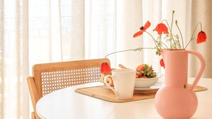 A round white table with a woven chair, a pink vase with red flowers, a white mug, and a plate with cookies and a small bowl of fruit. - rodakino - Seafront Apartment (Agia Effimia)
