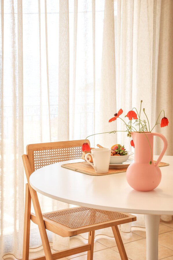 Uma mesa branca e redonda com uma cadeira de fibra natural, um vaso rosa com flores vermelhas, uma caneca branca e um prato com biscoitos e uma pequena tigela de frutas.