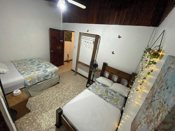 Une chambre avec deux lits simples, une porte en bois, un ventilateur de plafond et des guirlandes lumineuses.
