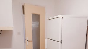 Pieni keittiö, jossa on valkoinen jääkaappi, metalliverkolla varustettu ovi ja laatoitettu lattia.