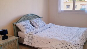 Siististi pedattu sänky kuviollisilla vuodevaatteilla, yöpöytä lampun kanssa ja ikkuna, josta avautuu näkymä rakennuksiin ja puihin.