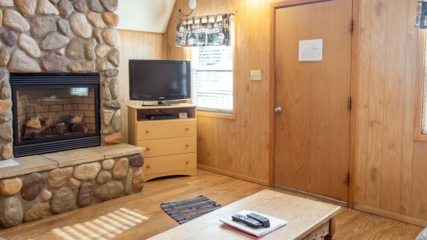 A cozy living room with a stone fireplace, a television on a wooden stand, and a wooden door with a sign. - Located on the river, facing the Mountains (Red River)
