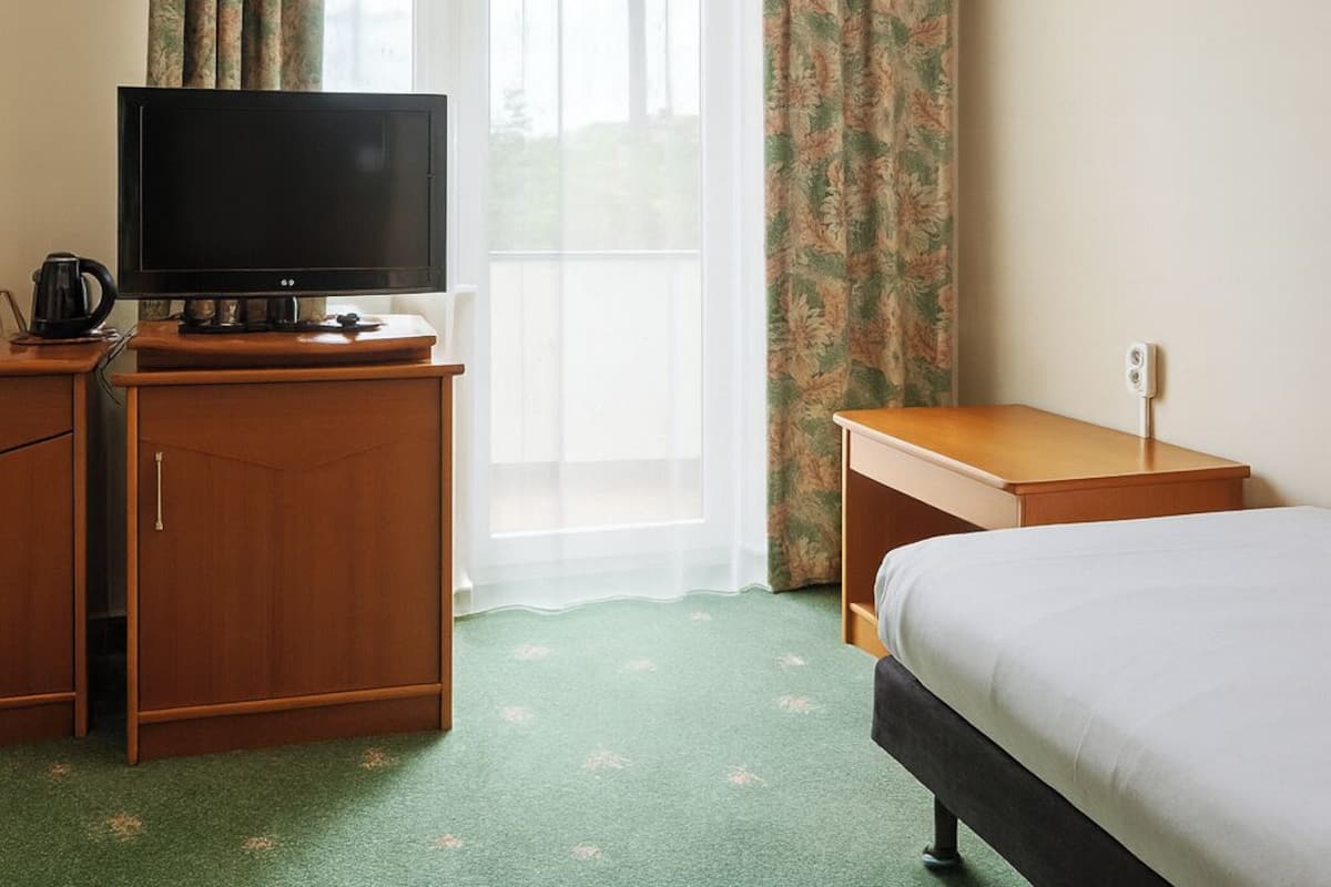 A hotel room with a television on a wooden stand, a bed, a small wooden table, and a window with curtains.
