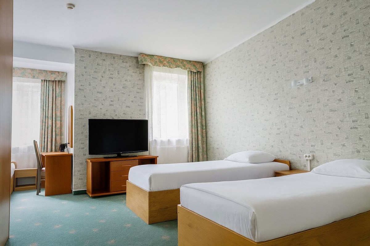 A hotel room with two single beds, a TV on a wooden stand, a desk with a chair, and a window with curtains.
