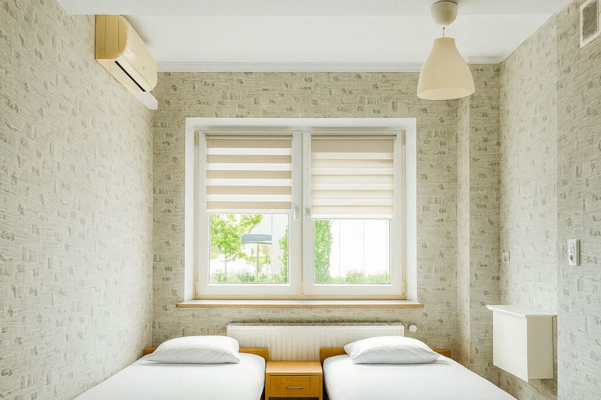 A room with two beds, a window with blinds, a ceiling fan, and a radiator.