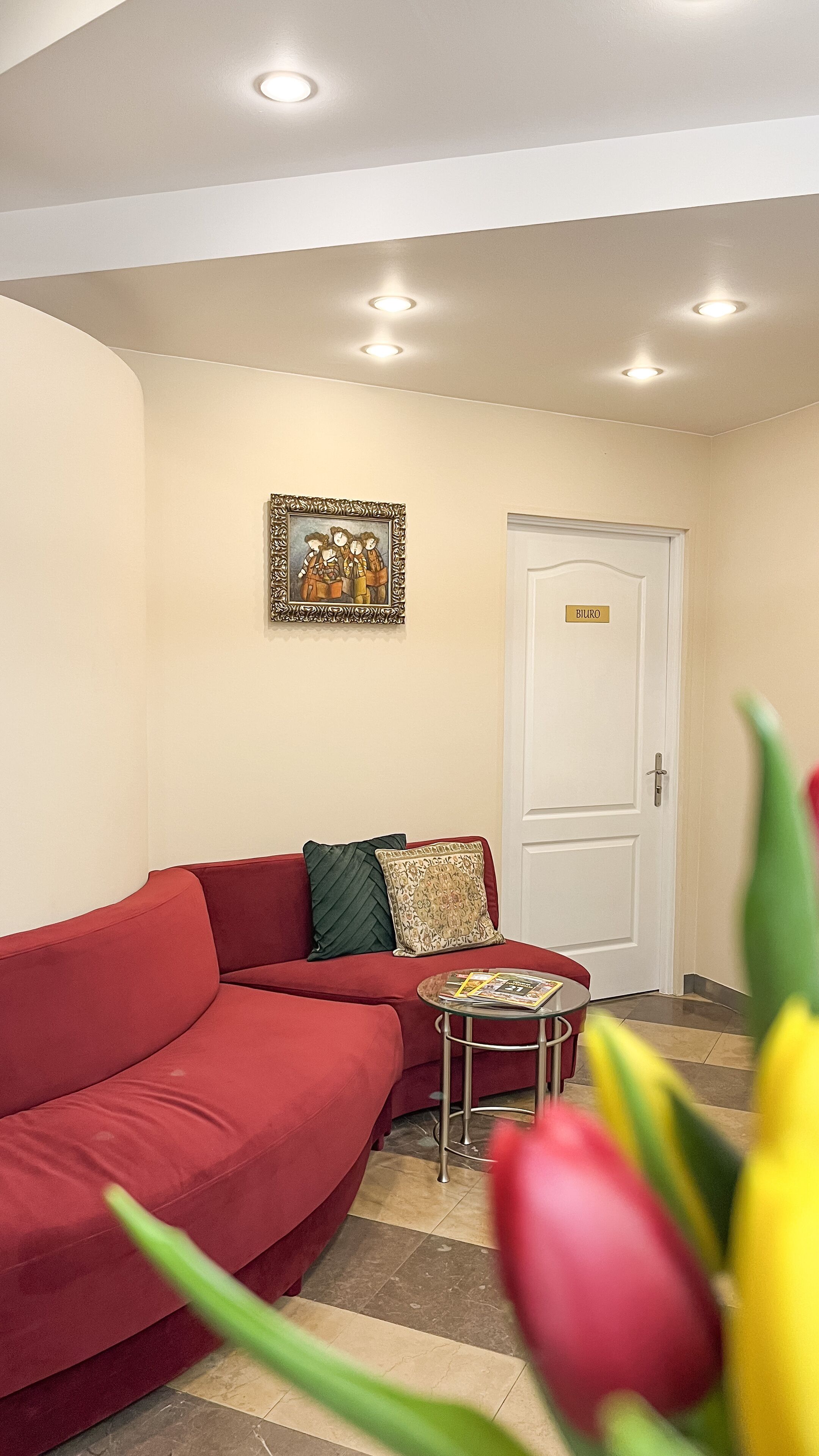 A room with a red sofa, a small round table with magazines, a door labeled "BIURO," and a framed picture on the wall.