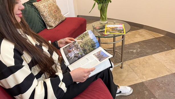Una persona leyendo una revista en un sofá rojo con un cojín estampado y una mesa de vidrio con una revista y un florero con flores.