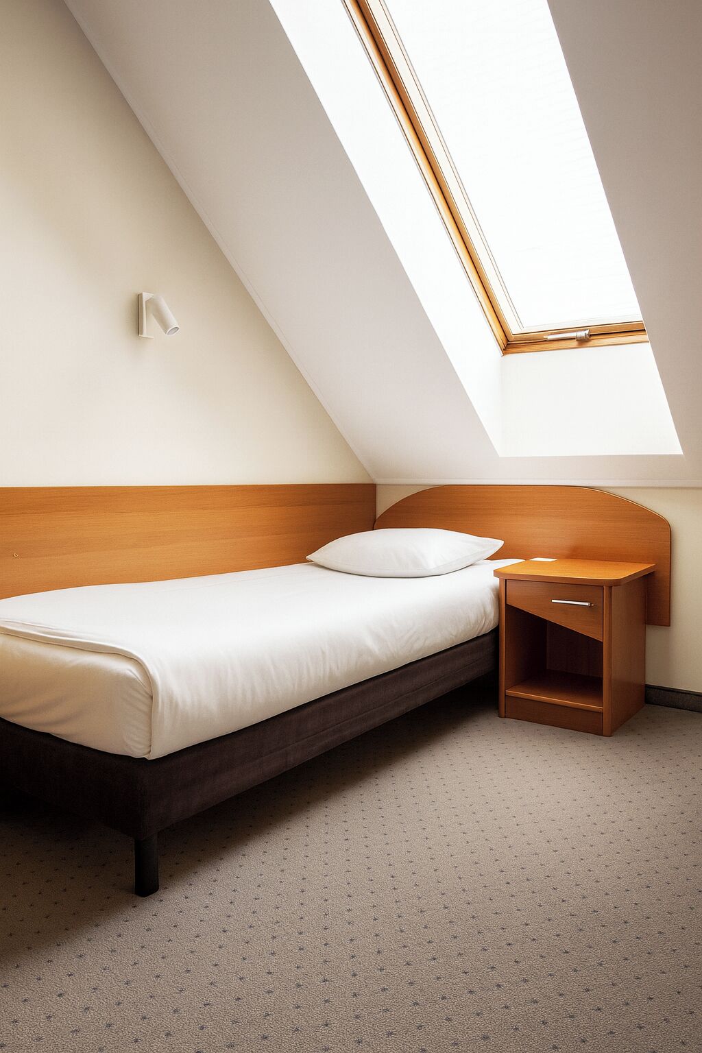 Een eenpersoonsbed met een houten hoofdbord, een nachtkastje met een lade, en een dakvenster erboven.