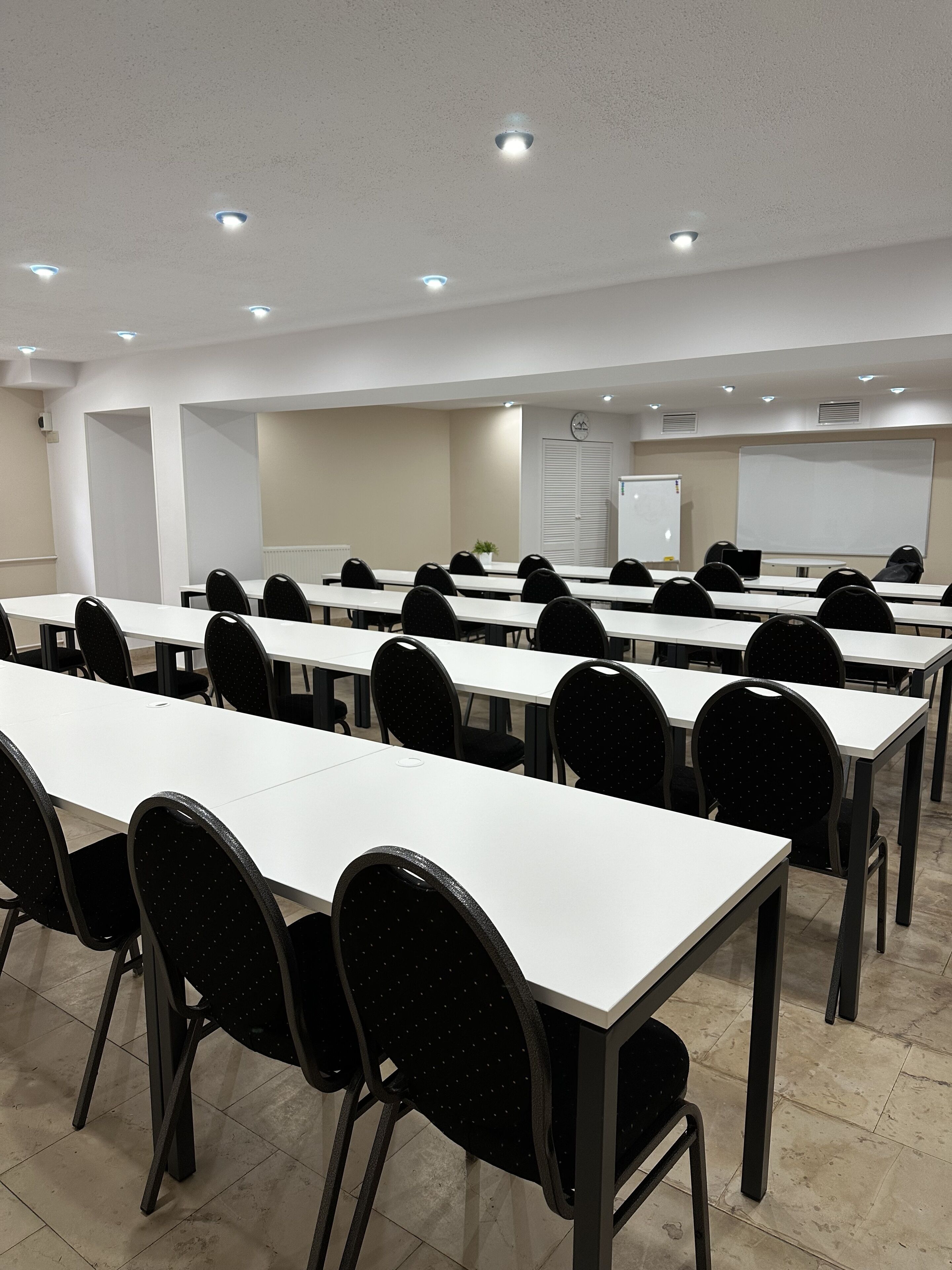 Un salón de conferencias con varias mesas blancas y sillas negras, equipado con una pizarra y una pantalla de proyector.