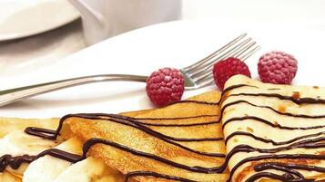 Un plato de crepas con glaseado de chocolate y rodajas de plátano, acompañado de frambuesas y una taza de café sobre una mesa blanca.