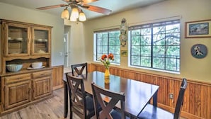 A dining area with a wooden table and chairs, a ceiling fan, and large windows with a view of trees.