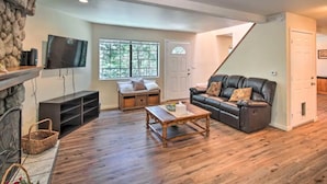A living room with a stone wall, a fireplace, a television, a sofa, a coffee table, and a window with a view of trees.