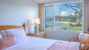 A hotel room with a bed, a nightstand with a lamp, a chair, and a large window offering a view of a beach and greenery.