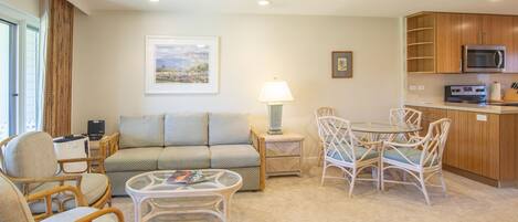 A living room with a sofa, chairs, a coffee table, and a dining table with chairs. There is a kitchen area with a microwave and cabinets.