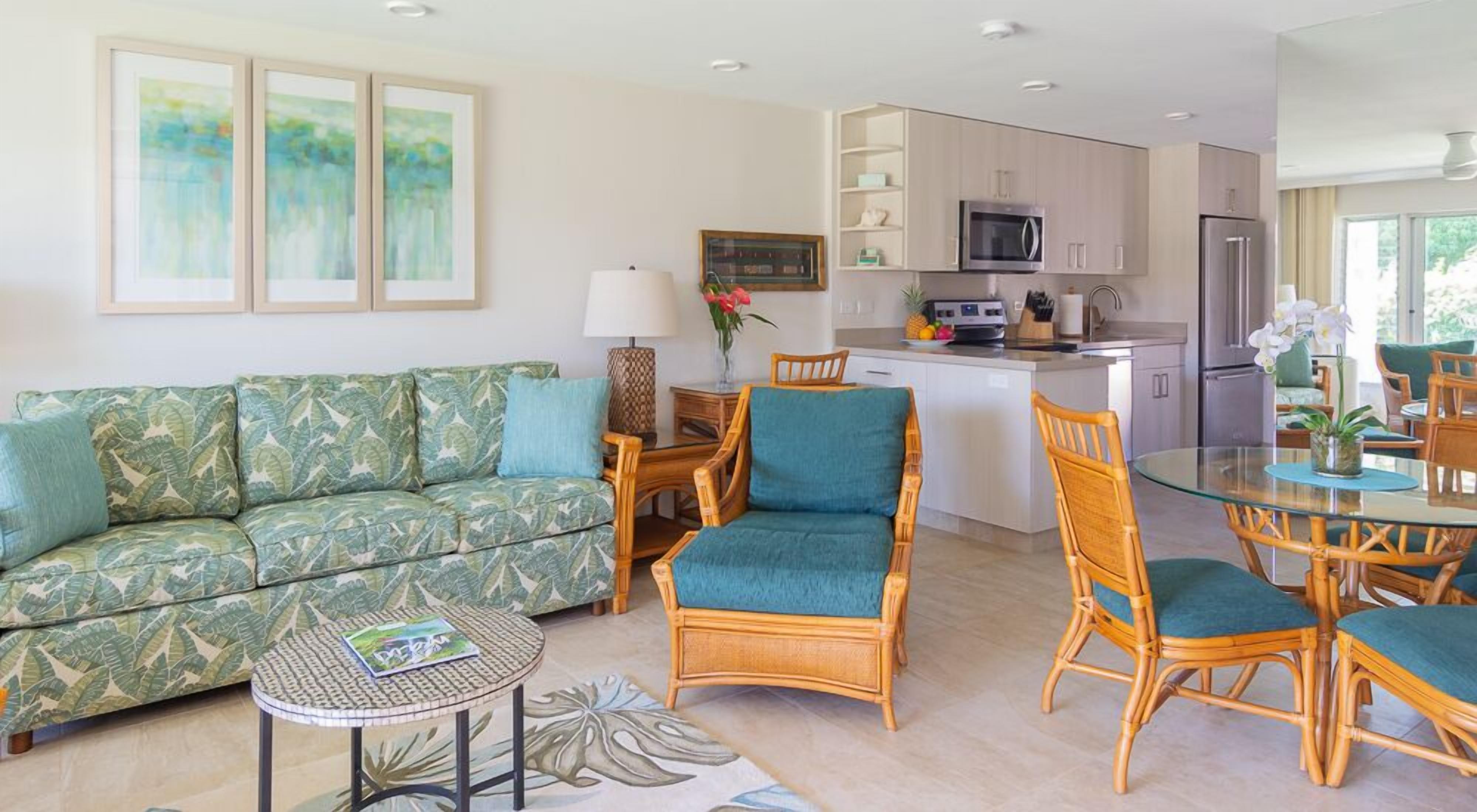 A modern living room with a green patterned sofa, a glass dining table, wicker chairs, and a kitchen with white cabinets and stainless steel appliances.