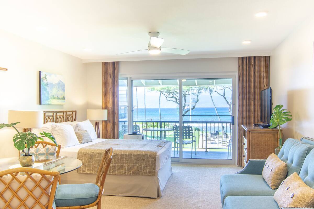 Une chambre d’hôtel avec un lit, un canapé, une table à manger et un téléviseur. Elle dispose d’un balcon avec vue sur l’océan.