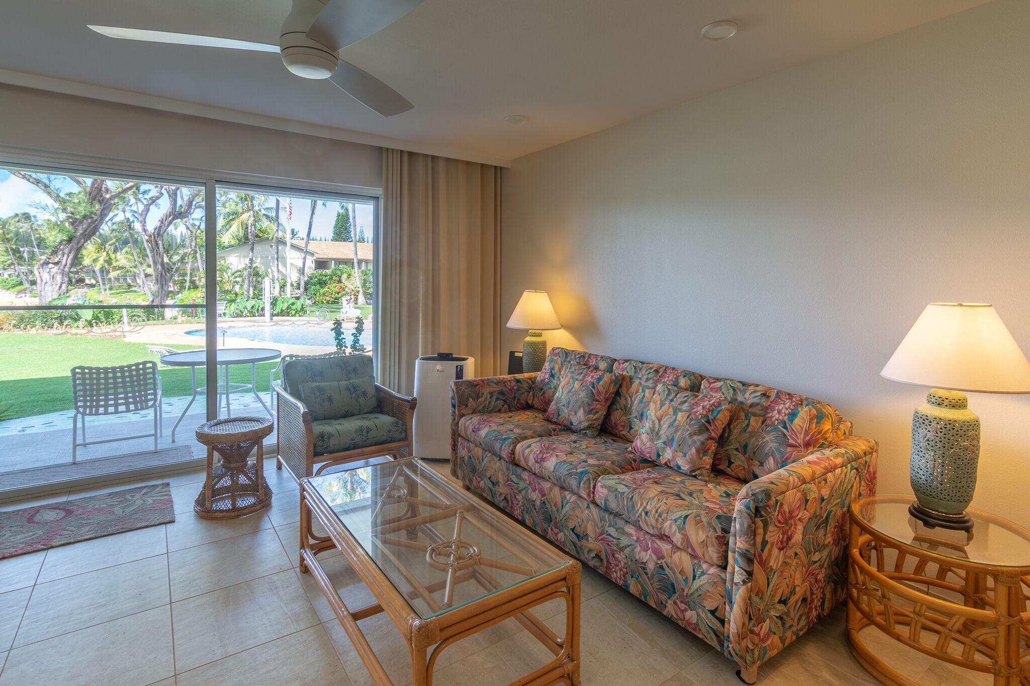 A living room with a glass coffee table, a floral sofa, wicker chairs, and a view of a pool and greenery outside.