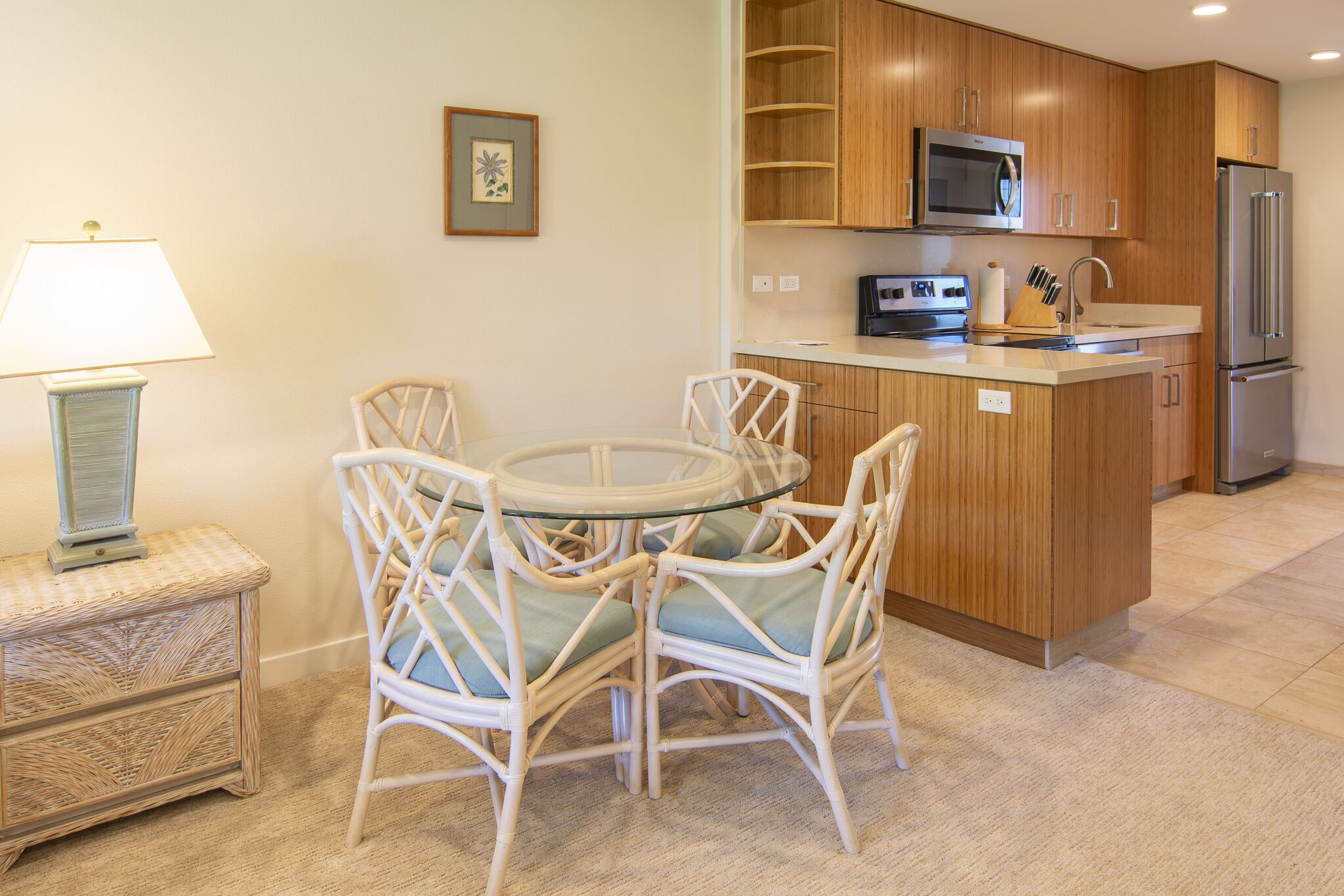 Una cocina con armarios de madera, microondas, refrigerador, una mesa de comedor con sillas y una mesita auxiliar con lámpara.