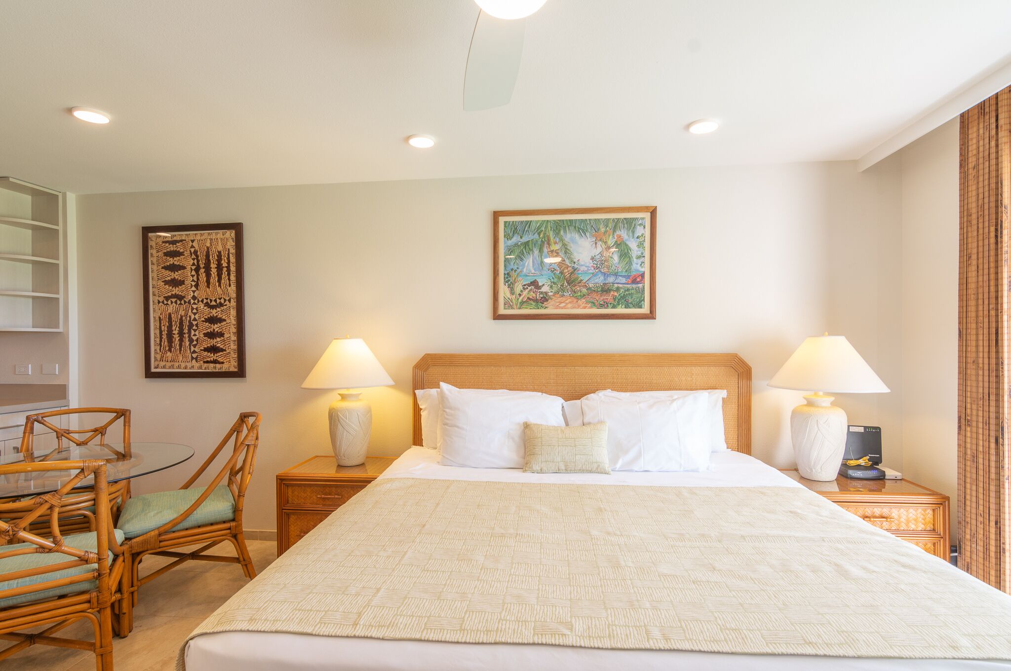 A hotel room with a large bed, two bedside tables with lamps, a wooden headboard, a painting on the wall, and a dining area with chairs and a table.